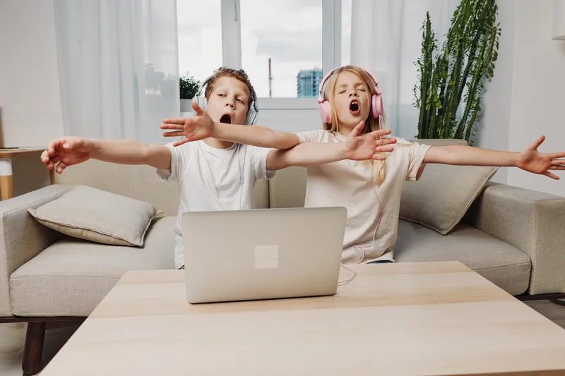 Two children with headphones sing loudly and stretch arms while sitting on couch at home with laptop on table. Concept of fun, music, and entertainment for kids.