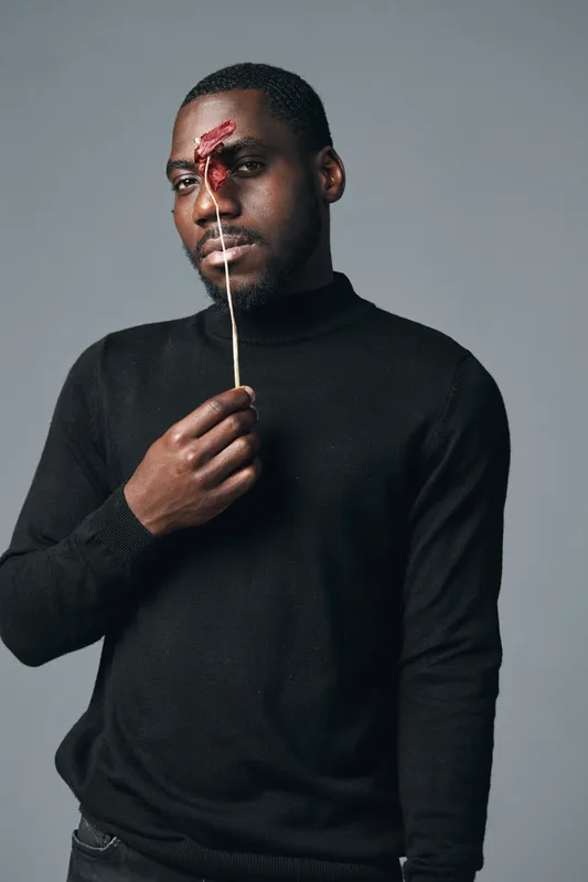 African man with dark skin wearing black sweater holding dried flower in front of face in creative conceptual visual style studio portrait on gray background.
