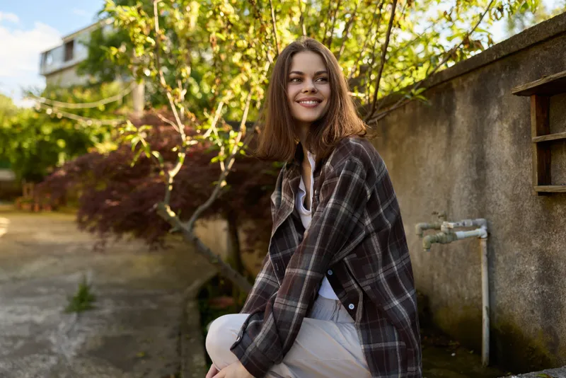 Cheerful woman outdoors, smiling softly, casual attire with a plaid shawl, sunlit garden setting, relaxed pose, gentle breeze, natural colors, warm and inviting mood, lifestyle portrait