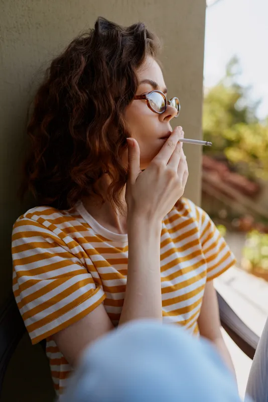 Young woman with curly hair in a striped shirt sits on a sunlit balcony, casually smoking a cigarette, looking out with calm confidence as a soft breeze brushes her hair.