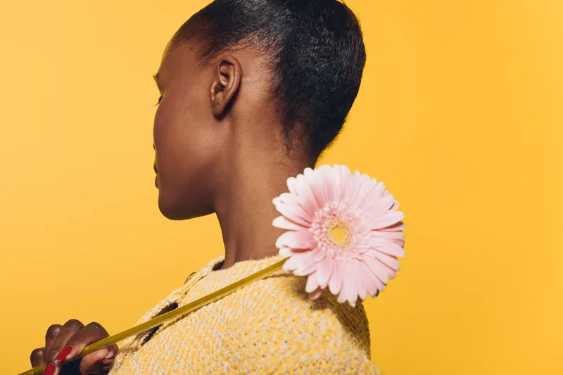 Profile of a young black woman with dark skin holding a pink flower on her shoulder against a yellow background. Concept of natural beauty, elegance, and floral aesthetics.