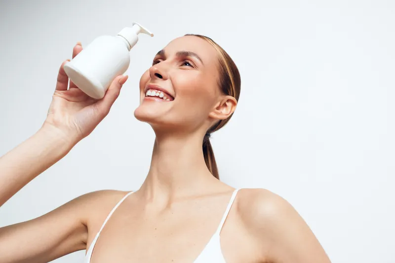 A smiling young woman in a white tank top holds a white pump bottle close to her cheek in a bright studio, illustrating a simple skincare routine and radiant confidence.