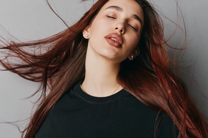 Young woman with long brown hair and closed eyes posing against gray background with natural makeup and relaxed expression in black top for beauty and fashion portrait.