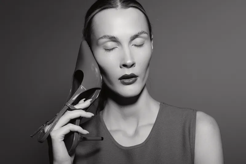 A pale white woman with closed eyes holds a high heel shoe near her face in a studio. Concept of fashion, elegance, monochrome, and creative portrait photography.