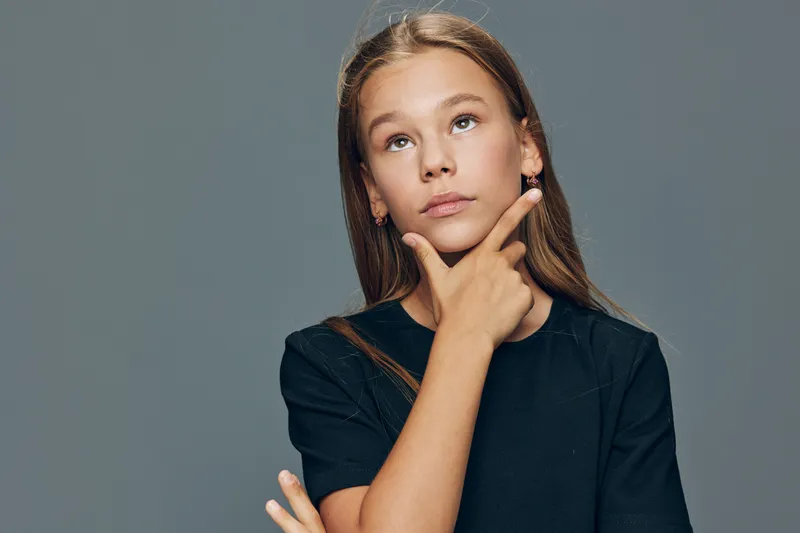 Girl thinking with hand on chin looking up wearing black outfit against plain background. Portrait of thoughtful child with long hair and pensive expression in studio lighting.