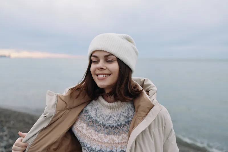 Woman portrait outdoors by the sea, wearing a knit sweater and light jacket, smiling with a gentle breeze and calm horizon, cold weather mood.