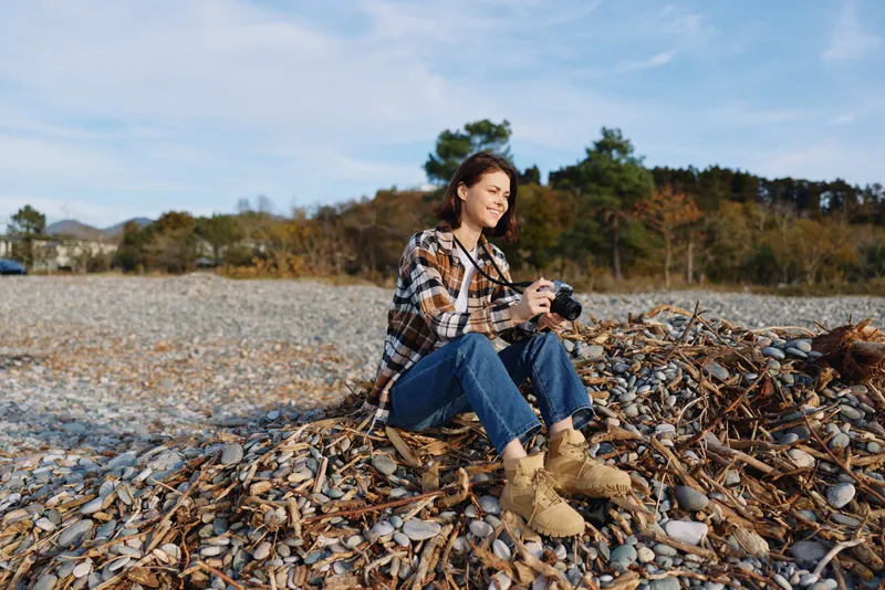 Woman with camera sitting on a pile of autumn leaves in a park, casual plaid shirt, jeans and boots, capturing photographs on a bright day.