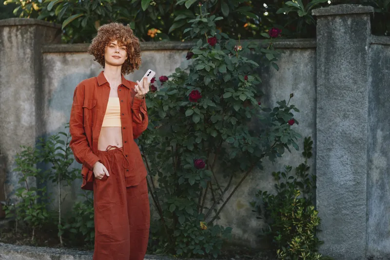 Smiling woman with curly hair wearing a red blazer and matching pants stands outdoors beside a stone wall and blooming roses, holding a smartphone. Bright natural daylight