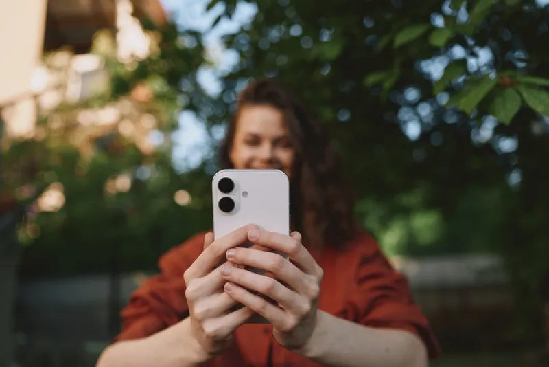 Smiling woman holding a smartphone in an outdoor setting for a casual selfie. Greenery in the background creates a fresh mood, emphasizing lifestyle, wellness, and everyday connection