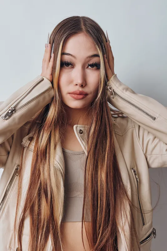 Young woman with long braids wearing beige jacket and crop top posing with hands on head looking at camera against plain wall background. Fashion, hairstyle and confident expression.