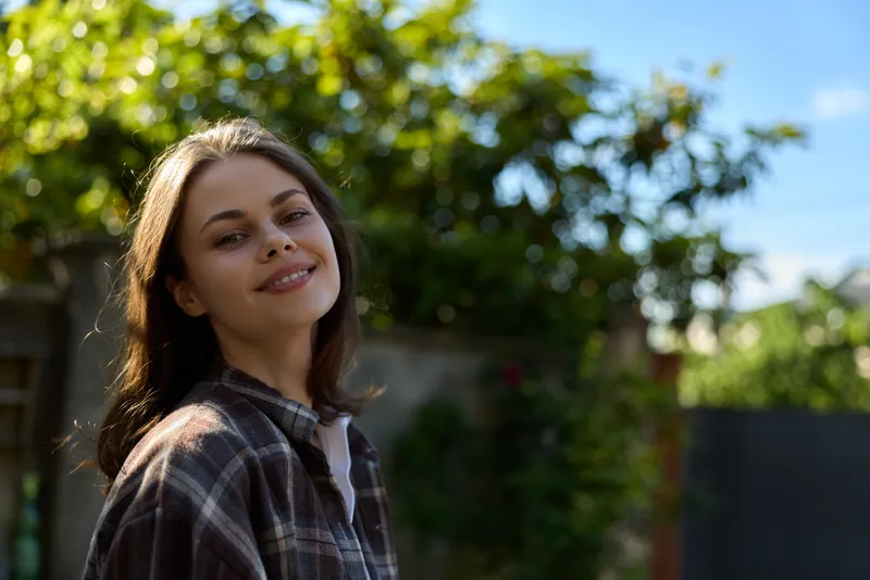 Casual relaxed woman outdoors wearing plaid shirt smiles softly in sunny garden setting, lifestyle portrait emphasizing natural light, carefree mood, and everyday confidence