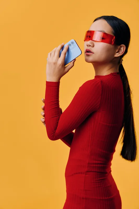 Woman in red dress and futuristic sunglasses holding smartphone near face on yellow background. Modern style, technology use, portrait with long dark hair and confident look.
