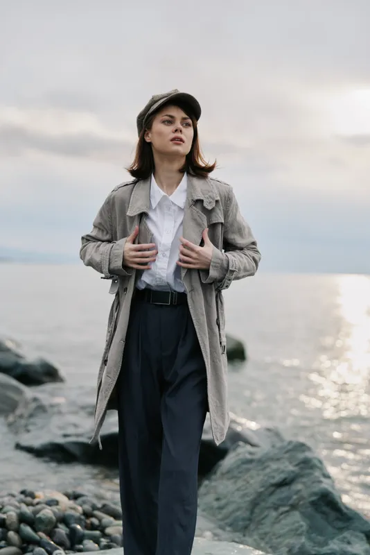 Young woman in a gray coat and cap stands on rocky shore near water, looking confidently into distance. Overcast sky and calm sea background create moody, contemplative atmosphere.