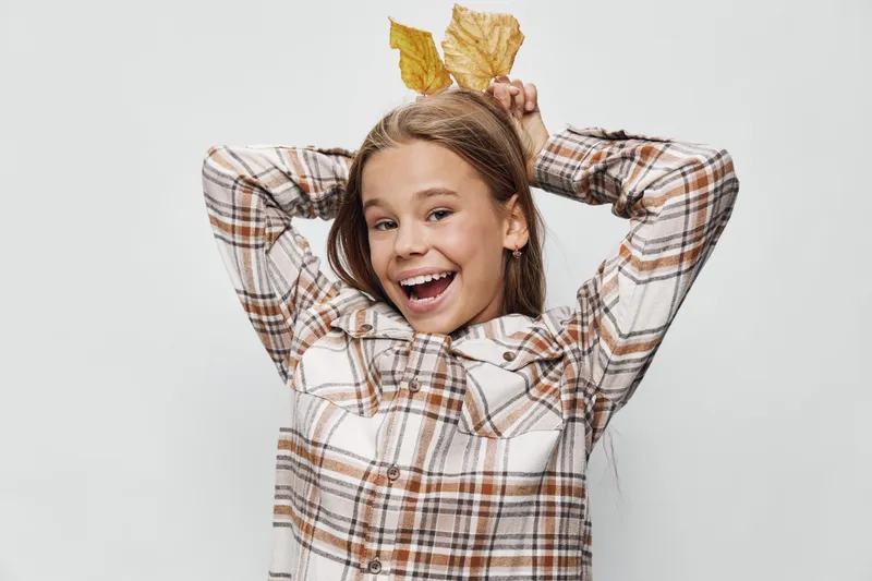 Girl with long hair smiling and posing with yellow autumn leaves on head wearing plaid shirt on white background. Happy child playing with seasonal nature elements.
