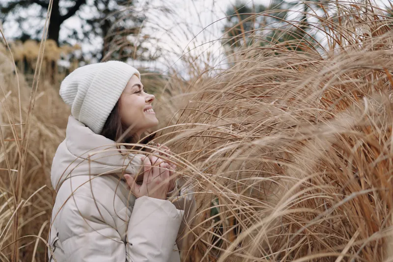 Woman in a winter coat and knit hat stands amid tall grasses, savoring a quiet outdoor moment as soft reeds blur in the background, depicting calm nature and intimate seasonal mood.