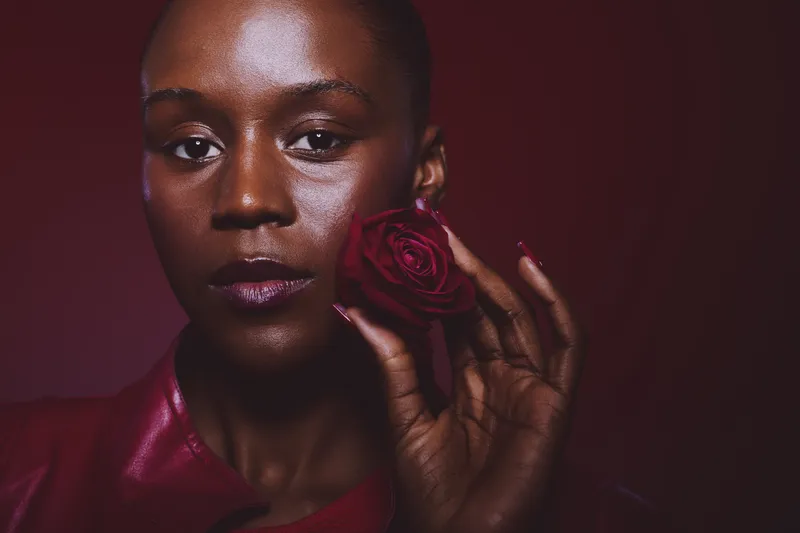 Close-up of a dark skinned African woman with shaved head holding a red rose near her face against a dark red background. Concept of beauty, elegance, and natural skincare.