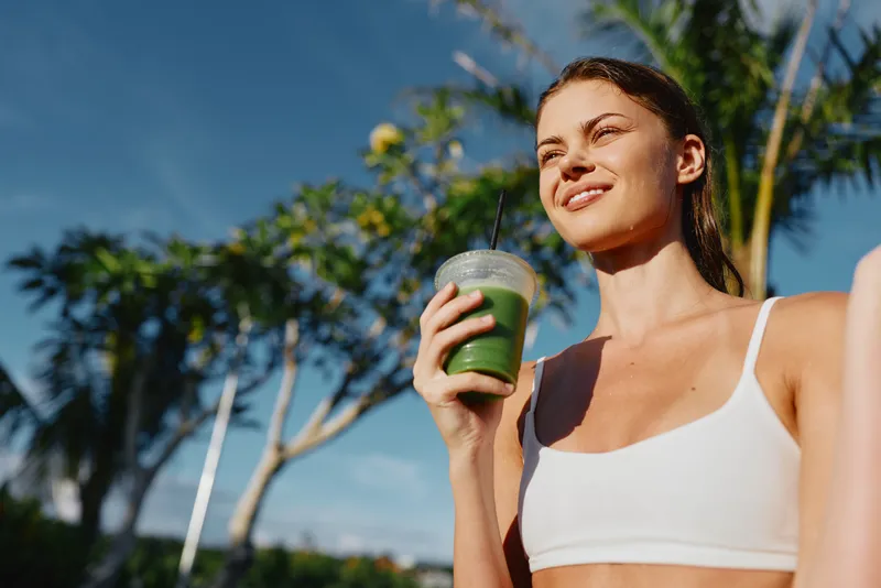 Cheerful woman outdoors, wearing a white sports bra, sipping a green smoothie, sunlit moment, relaxed posture, tropical trees and blue sky in the background, natural colors, casual lifestyle