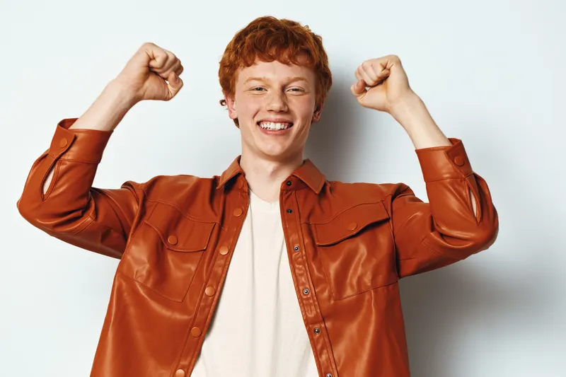Young man with red hair smiling and raising arms showing excitement and success in casual brown shirt against white background. Portrait, happiness and positive emotion.