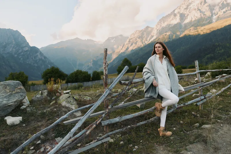 Woman in a cozy white outfit leans on a rustic wooden fence in a mountain valley, capturing a calm lifestyle amid stunning scenery and fresh air.