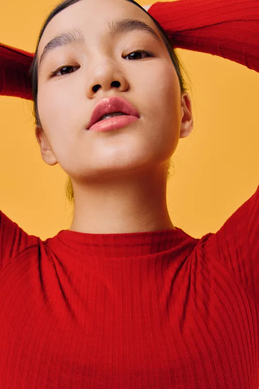Woman portrait looking down with confident expression wearing red sweater on yellow background. Young female model with smooth skin and natural makeup in studio lighting.