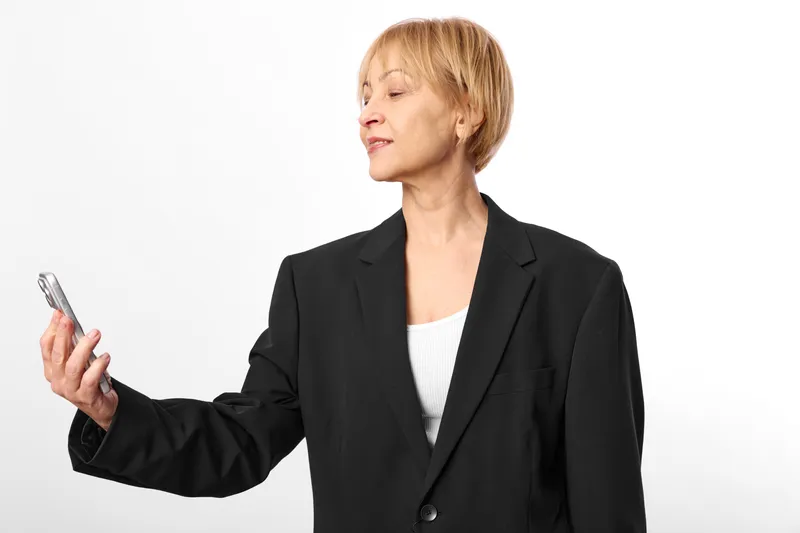 Woman in black blazer holding smartphone in studio white background, business communication and technology concept, portrait of mature professional checking mobile content, soft neutral light