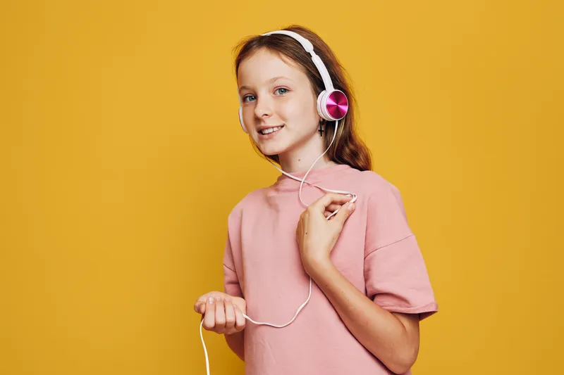 Smiling girl with red hair wearing pink headphones and a light pink shirt on a vibrant yellow background. Concept of music enjoyment and youth lifestyle.