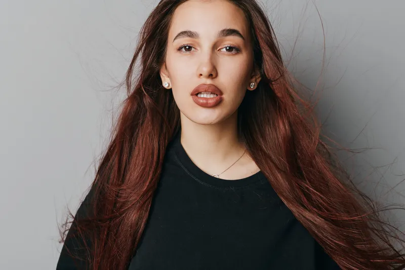Young woman with long brown hair wearing black shirt posing against gray background. Natural makeup, earrings, and casual style with loose hair for beauty portrait and fashion concept.