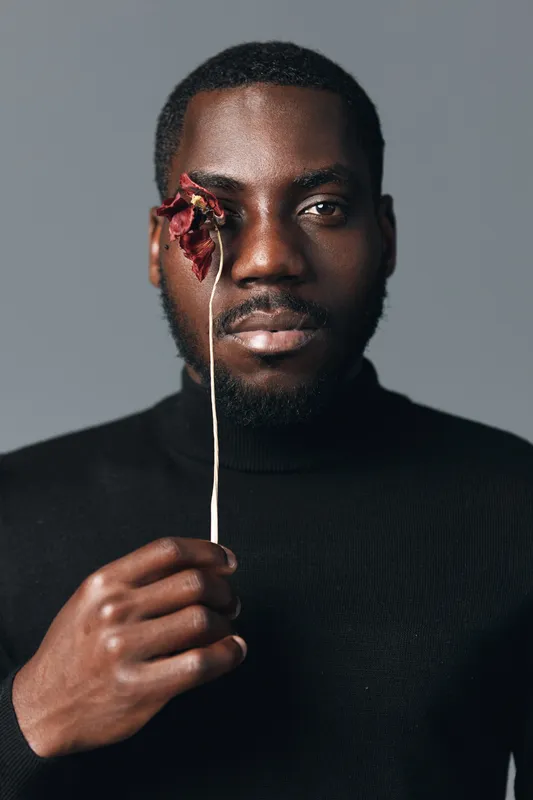 African man with dark skin holding dried flower in front of face wearing black turtleneck sweater studio portrait creative conceptual visual style isolated on gray background.