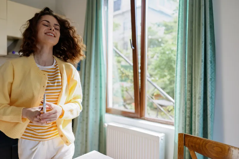 Smiling young woman wearing a yellow cardigan and striped shirt stands by a sunlit window in a cozy home, enjoying a carefree, uplifting moment of everyday life