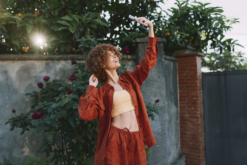 A young woman with curly hair dances in a sunlit backyard, wearing a mustard crop top and rust suit while smiling and raising her arms against a leafy garden backdrop