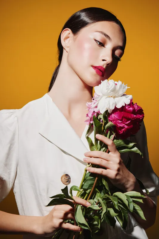 Elegant woman in white blazer holds vivid flowers against bright yellow background. Her glowing makeup and graceful pose express beauty, fashion, and creative energy.