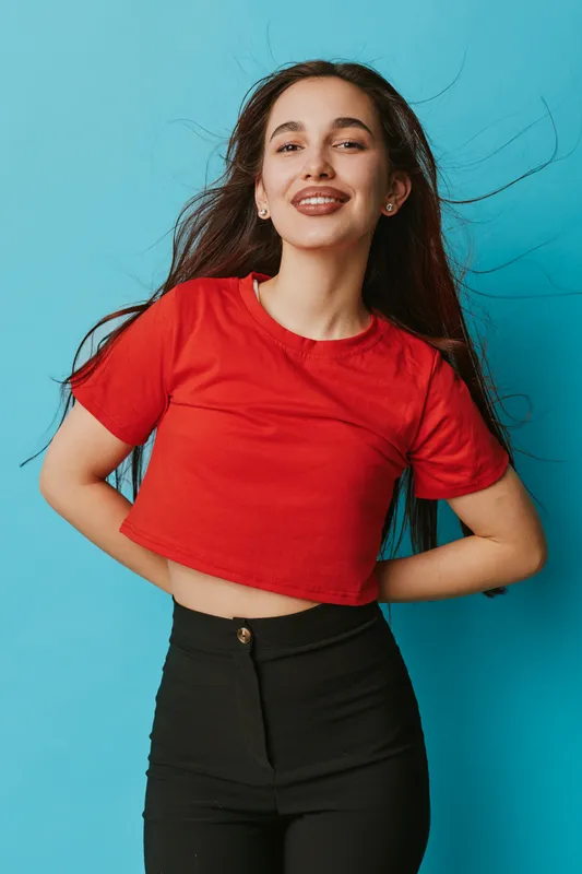 Young woman wearing red crop top and black pants posing with hands behind back against blue background, smiling confidently with long hair flowing in wind.