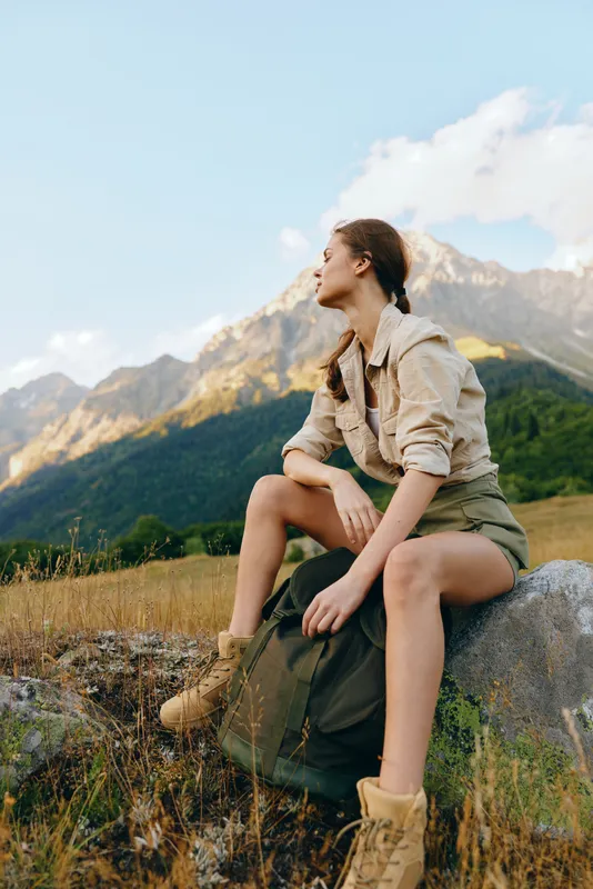 Young woman with braided hair sits on a rock in hiking boots and casual jacket while enjoying a mountain landscape during a sunny day outdoors.