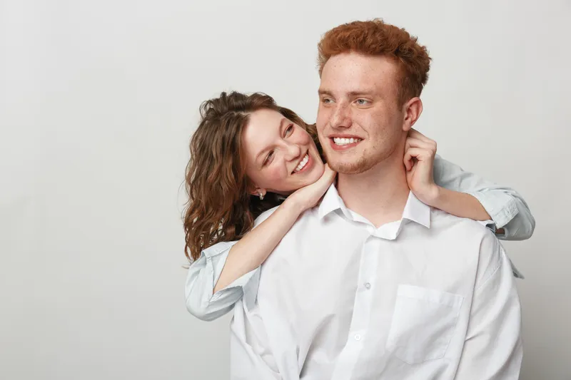 Happy young couple in casual white shirts embraces and smiles against plain background. The woman hugs the man from behind, expressing affection and joy in a loving moment.