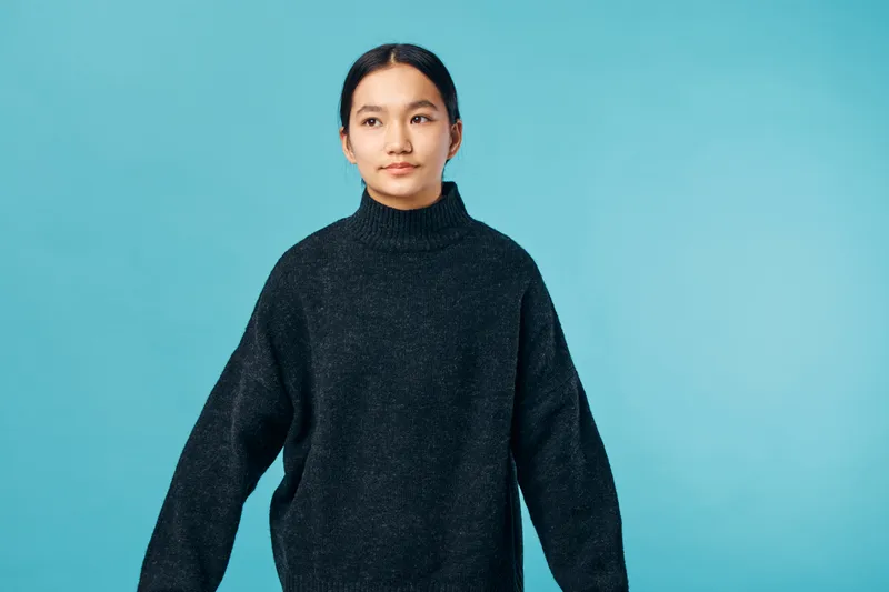 Portrait of young woman in black sweater standing against blue background looking sideways with calm expression casual style and minimalistic setting in studio environment.