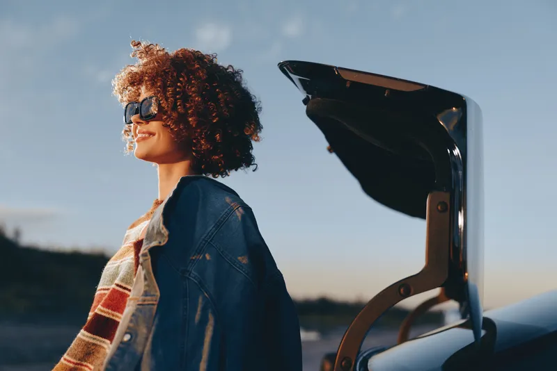 Woman outdoors lifestyle with curly hair wearing denim jacket, standing by water at sunset, smiling in soft breeze, using a telescope on a boat, warm golden light, evening wide sky