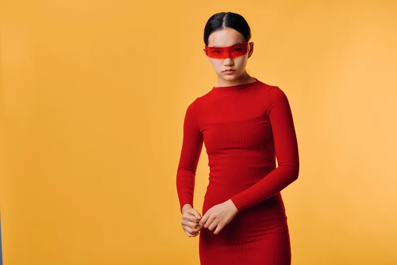 Woman in red dress and futuristic glasses posing in studio with orange background. Fashion model stands confidently showing modern style and minimalistic aesthetic in portrait.