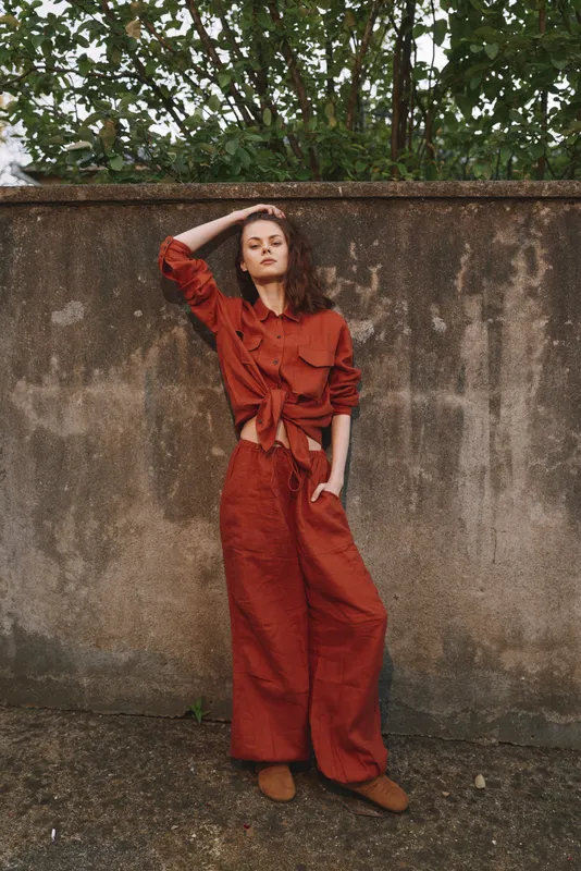 Young woman in a rust red two piece outfit stands against a weathered wall, hands relaxed and gaze confident, conveying casual street style and poised mood