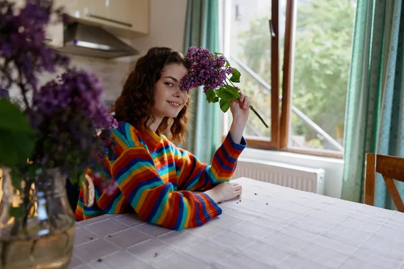 A woman with curly brunette hair sits at a sunlit dining table in a cozy indoor kitchen, holding a purple bouquet and smiling, creating a warm home moment.
