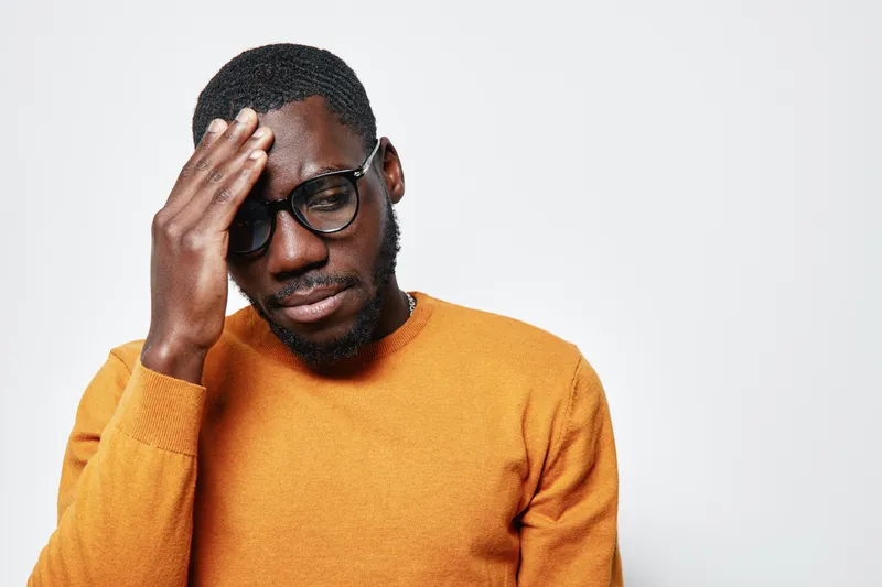 Black man with dark skin wearing glasses and orange sweater touching forehead with closed eyes in stress or headache on white background. African ethnicity, casual clothing and facial expression.