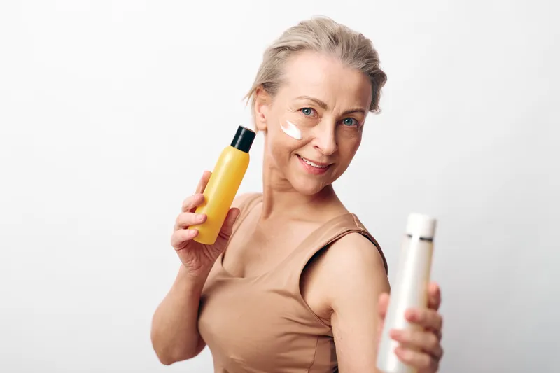 Smiling middle aged woman with clear skin applies cream on her cheek holding two skincare bottles. Concept of natural beauty, skincare routine, and healthy skin care products.