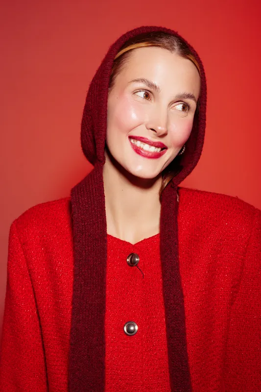 Smiling woman in red sweater and hood looks away on red background. Bright makeup and glossy skin create vibrant fashion portrait with warm and joyful mood.