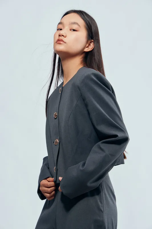 Young woman wearing dark blazer standing confidently with hands near waist, looking upwards against plain light background. Female model with long hair and professional outfit pose in studio.