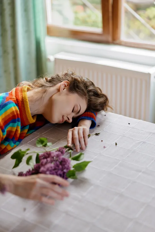 Young woman rests her head on a table beside a bouquet indoors, wearing a colorful striped sweater, conveying quiet exhaustion, tenderness, and a moment of vulnerability in soft daylight.