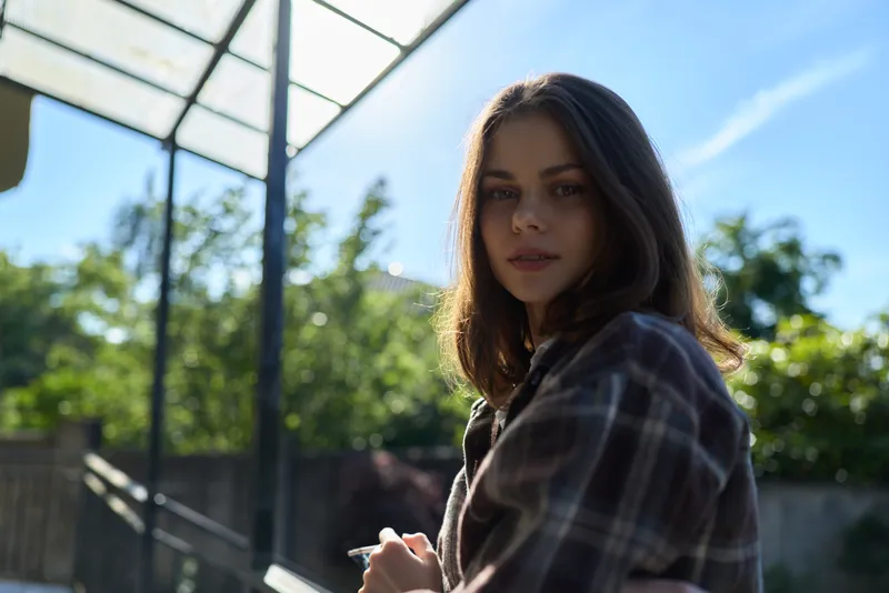 Casual outdoor portrait of a confident woman in plaid shirt, smiling under bright sunlight, surrounded by greenery, conveying a relaxed lifestyle and natural beauty