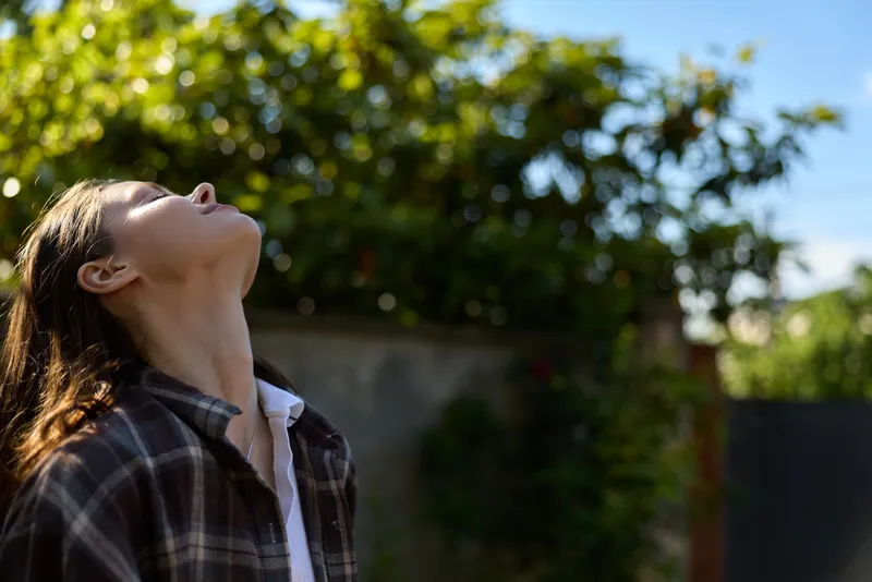 Young woman tilts her head back to enjoy the sunlight in a casual outdoor garden scene, wearing a plaid shirt and embracing fresh air, calm and relaxed mood
