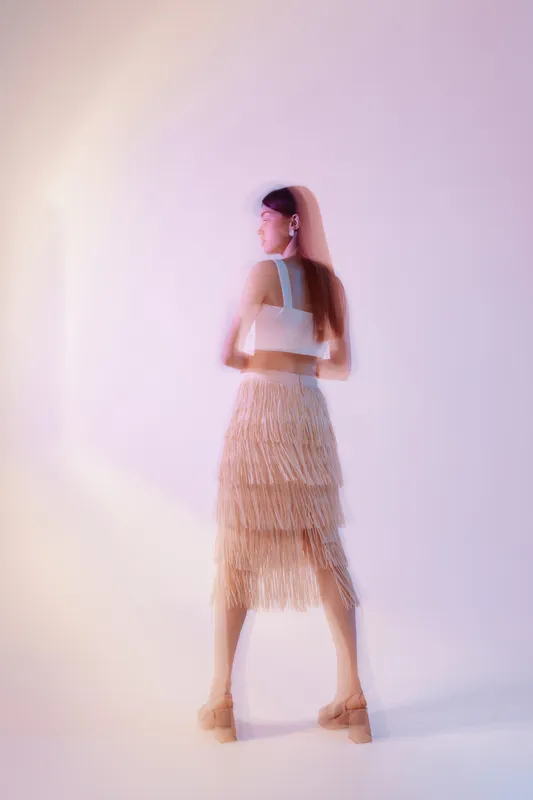 Young woman in a stylish fringed skirt and sleeveless top poses with her back turned. Soft lighting and pastel background create a modern fashion beauty concept.