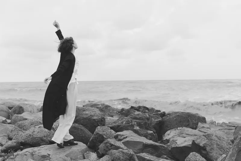 Black and white image of a couple dancing on a rocky shore, wind in coats, embrace mid motion, waves crashing nearby, moody seaside scene conveying freedom, movement and shared escape