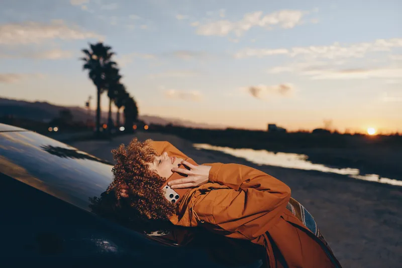 Woman lifestyle beach sunset portrait nature ocean waves outdoors calm Rust coat woman reclines on a rock at sunset on the ocean shoreline, golden light, curly hair with floral accents, relaxed