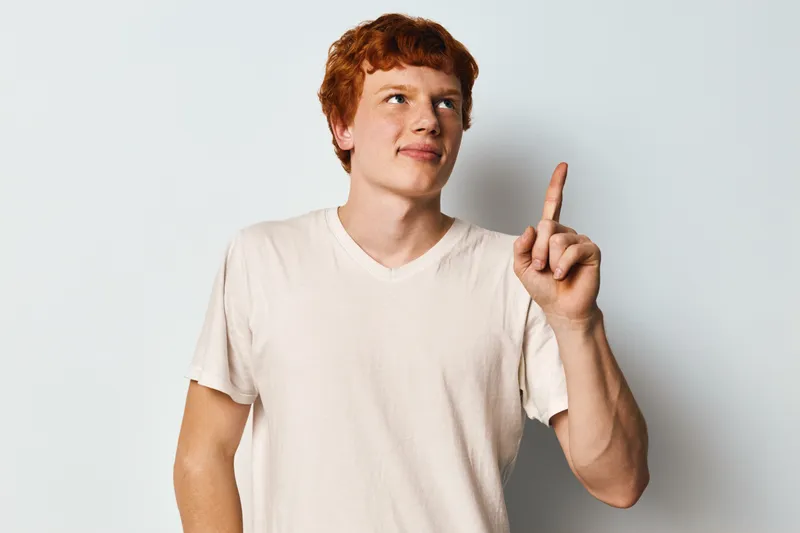 Young man with red hair and freckles wearing white t shirt pointing finger up while looking aside standing against white background. Casual people and gesture concept for attention or idea.